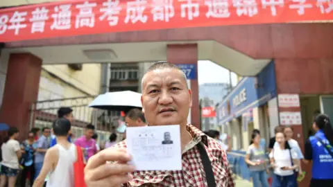 GETTY IMAGES Liang Shi holds his ID up in front of a 2018 National College Entrance Examination building on 07 June, 2018 in Chengdu, Sichuan, China.