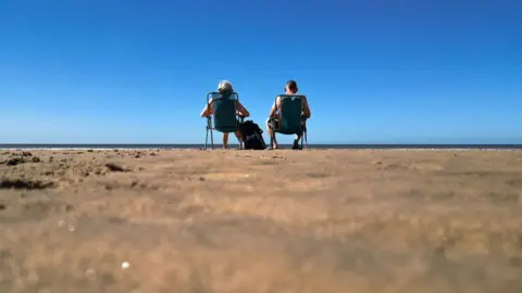 Christopher Furlong/Getty Images A man and a woman sit on deck chairs under blue skies on Blackpool beach