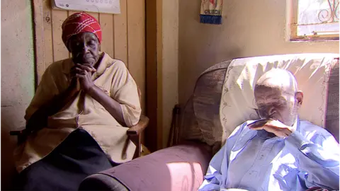 BBC Vesta (L) and Teddy are a married couple. They sit on chairs in their home in Zimbabwe.