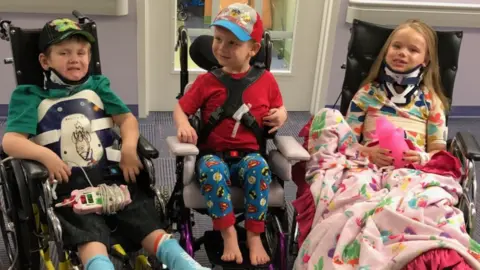 Teresa Burrell Three children sit together. All are in wheelchairs, with various casts and braces.