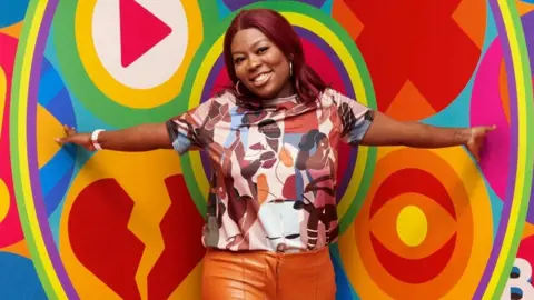 Big Brother contestant Trish wearing orange trousers and a colourful top with people on it, in front of a multicoloured background