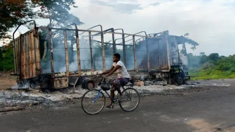 Getty Images A biker passes by a burned truck on the side of the road in a village near Beoumi on May 17, 2019 after violence erupted between members of the Baoule local community and northern ethnic group Dioula people. Fighting erupted between people of the Baoule and Dioula tribes Wednesday at Beoumi, a town in the centre of the country, after an altercation between a taxi driver and a motorbike taxi operator at a taxi rank, said witness Innocent Koffi, a local farmer. The clashes caused nine deads and 84 injured people including six gendarmes and several houses and vehicles were set ablaze.