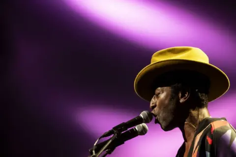 EPA Keziah Jones sings into a microphone. His eyes are closed and he wears a yellow, wide-brimmed hat tipped forward. The background is a vivid purple.