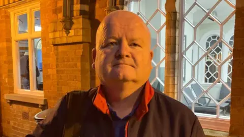 Alex Pope/BBC Man with bald head standing at a railway station