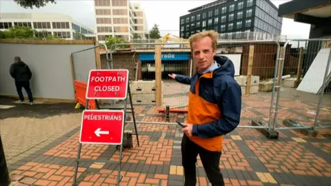 BBC reporter Dan O'Brien next to red signs with white lettering reading footpath closed and pedestrians with an arrow pointing left