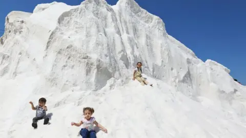 Getty Images Three children sitting on a salt mound. It is bright white, against a backdrop of blue skies.
