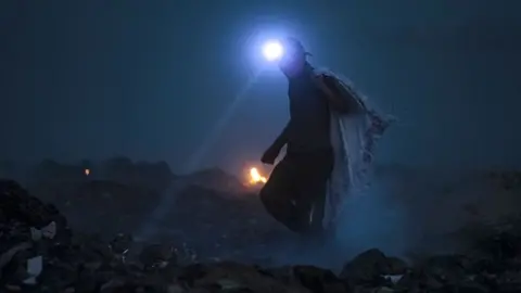 A man equipped with a torch scans for recyclable material at a landfill