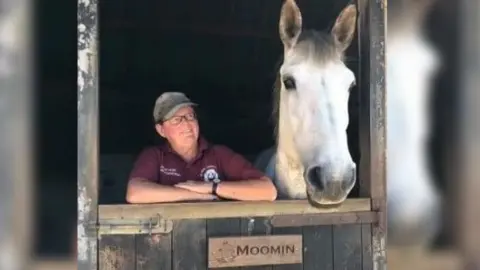 Wiltshire Police A woman in a burgundy top and a cap standing next to a white stallion