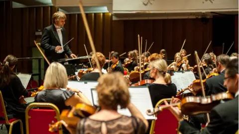 Chris Cooper Kiril Karabits in formal dress and holding a conductors baton is standing facing an orchestra and conducting them while they play