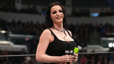 AEW Saraya smiles at the camera. She's in the middle of a wrestling ring, holding a microphone and wearing a vest top and chunky chain necklace