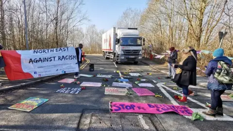 Laura Lee Daly Flowers and placards laid on road with campaigners stood around outside centre