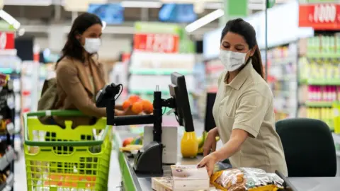 Getty Images A woman shopping for food wears a face mask, as does the cashier