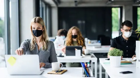 Getty Images Young office workers masked up look at their laptops in an office setting