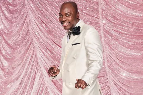 Ray Burmiston/BBC Eddie Kadi wearing a white tuxedo jacket and black trousers