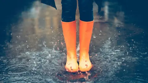 Getty Images Close-up of woman in orange rubber boots jumping on the puddle