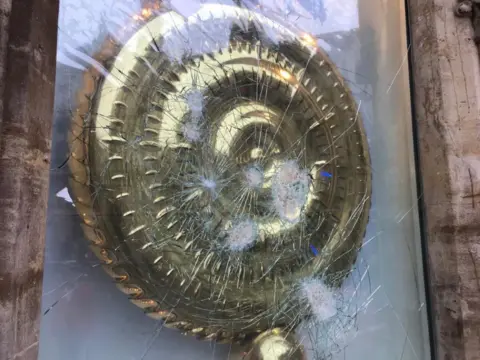 Orla Moore/BBC The gold clock is behind glass which has been partially blown out but not entirely smashed. It has large cracks across the glass from the points of impact.