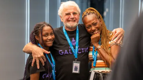 Virgin Galactic Anastatia (Ana) Mayers, Jon Goodwin and Keisha Schahaff hug after their voyage to space. Jon has his arms around Ana, on his left, and Keisha on his right. Ana, an 18-year-old black woman, has her hair styled in long braids tied back in a pony tail. She has piercings on the bridge of her nose, her septum and her lip and smiles at the camera. She wears a black T-shirt with 'Galactic' written in purple on it - Jon is wearing the same T-shirt and looks emotional while smiling at the camera. Jon is a white man in his 80s and has short white hair and a white beard and wears rimless glasses. To his right is Keisha, a black woman in her 40s, who is also smiling and looking emotional. Keisha has her hair, dyed blonde, in braids down to her middle tied back behind her head. She wears a white, brown, green and orange striped T-shirt and holds her right hand to her face.