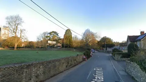 Google The B3081 Station Road In Bruton. There are houses to the right and a wall and field to the left of the road. In the distance, some roadwork signs and a line of cars can be seen.