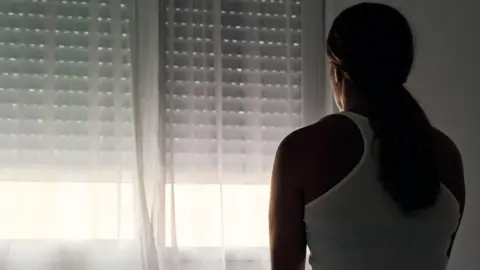 Rear view of an unrecognisable abused woman sitting on her bed looking out the window