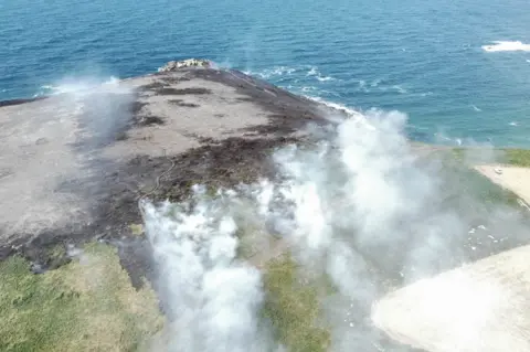 National Trust Aerial view of smoke and burnt patch of grass on cliff top next to sea