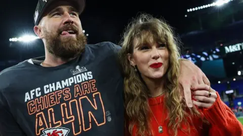 Getty Images Travis Kelce and Taylor Swift, he is wearing American Football uniform and a hat and has his arm around her - she is wearing a red top and smiling