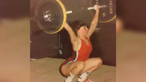 Trish Tenn A photo from Trish taken in the 90s where she is squatting whilst holding a barbell above her head