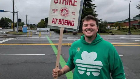 Un Marc Cisneros sorridente che indossa una felpa con cappuccio verde con un trifoglio bianco, in piedi di fronte a una strada deserta fuori dal Boeing con in mano un cartello di sciopero