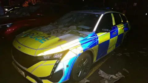 Cambridgeshire Police Another view of the police car from the side with smashed ceramic tiles on its front windscreen and bonnet. The windscreen has been smashed by the tiles. Smashed tiles can be seen on the ground around the vehicle.