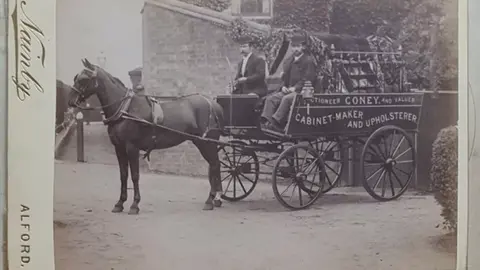 Paul Andersen A horse and cart, with two male passengers, in a black and white photo, possibly dating from the late 19th Century. The cart carries the words "Coney, cabinet-maker and upholsterer".