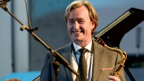 A man in a grey suit and white shirt holding a saxophone 