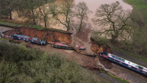 PA Media An aerial view of the collapse - showing water flowing into a nearby field. There is a large hole in a canal and the section is dry and waterless. Two boats are in the hole, another on dry, higher ground hovering over the edge of the hole.
