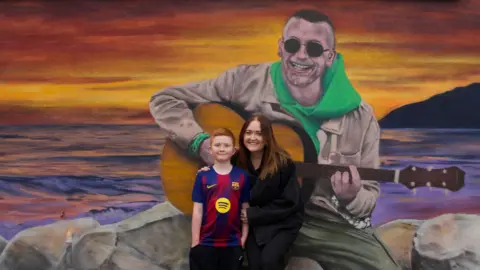 A mural of a man sat on rocks, with the sunset behind, playing a guitar. In front in a boy with ginger hair wearing a blue and purple Barcelona football shirt. Next to him is a woman in a black top with long brown hair