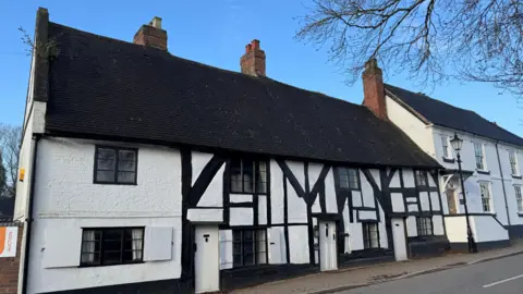 A timber-framed, black and white building. There is another, taller, white building next to it and they are both set against the backdrop of a blue sky.