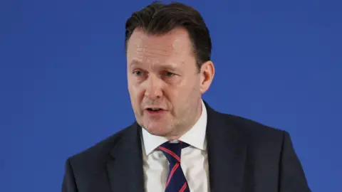 PA Media Russell Findlay has short dark hair and wears a dark suit and white shirt with a navy and red striped tie. He is mid speech and in front of a blue background