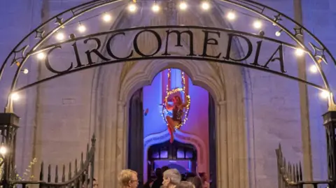 Circomedia The entrance to the Portland Square Circomedia building which has its name in an arch in iron above its metal gates. A performer can be seen in its foyer on an aerial hoop decorated with fairly lights as people file into the building.