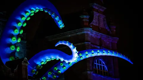 Lancashire County Council Blue inflatable tentacles from a giant inflatable creature attached to a tall building in Morecambe at night.