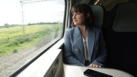 Rachel Reeves looking out of a train window. She is seated and looking to her right. She has dark hair and is wearing a pale blue jacket and white blouse. A mobile phone is placed on the table in front of her. 