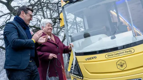 Transport for Greater Manchester Andy Burnham and Sharon Goodman. Andy has grey/black hair and glasses, He wears a navy blue jacket, jeans and a scarf. Sharon wears a wine coloured thick overcoat and a scarf. She has grey hair and a flower in her lapel. 