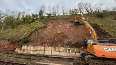 Network Rail An orange vehicle parked on a railway line next to a visibly damaged embankment. Concrete blocks have been placed at the bottom of the hill. The vehicle's arm is extended towards the embankment.