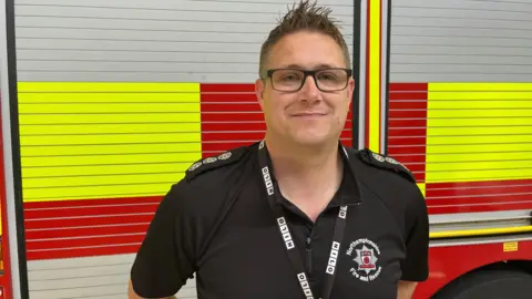 Nick Gayton in a black polo shirt with black hair and glasses, standing in front of a fire engine.