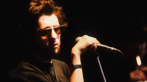 Shane MacGowan is pictured in semi-silhouette against a black background. He is wearing tinted sunglasses and holding a microphone, while preparing to sing with the Pogues