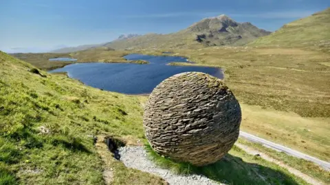 Michael O'Kane A circular sculpture made of individual rocks on a hill overlooking a loch and grassy hills 