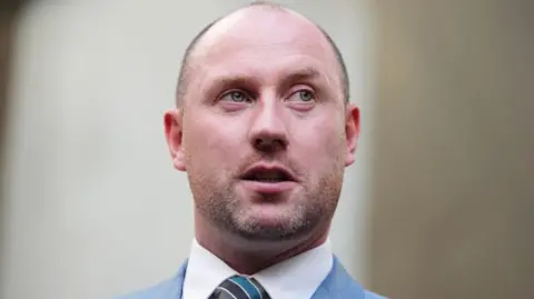 PA Media Neil Gray, a bald man in a white shirt and blue suit, looks to his left