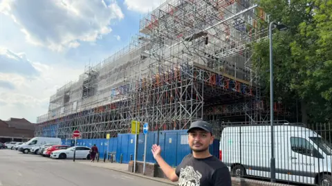 Alperton councillor Anton Georgiou in front of the building which is fully hidden behind scaffolding. Georgiou is wearing a black baseball cap and a black T-Shirt with a white logo and writing on it.
