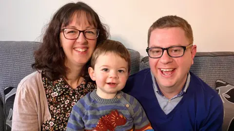 Lady with brown hair, glasses and a flowery dress and beige cardigan smiling. Man with short sandy hair, glasses and checked shirt and blue V-necked sweater smiling. Boy toddler sits between them on the sofa with stripy jumper with dinosaur on it. 