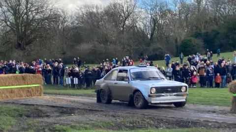 An old, dirty white Ford car is driving down a road outside the town centre. There are trees in the background. Spectators are standing behind a cordon. There are hay bales on the left.