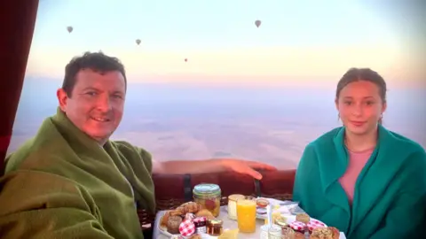 Hollinson family Michael and Darcy sat together in a hot air balloon. There is a small, round table with food and drinks between them and they're both wearing blankets over their shoulders. They're smiling at the camera. 