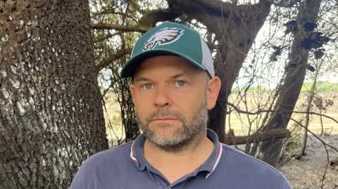 John Devine/BBC A man - Chris Milligan-Smith - looking straight at the camera with a neutral expression. He is wearing a navy polo shirt and a green hat. He is standing in front of a tree and fields.