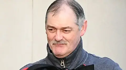 Daniel McClean. He has grey hair and a moustache, brown eyes and wearing a grey zip up jacket.