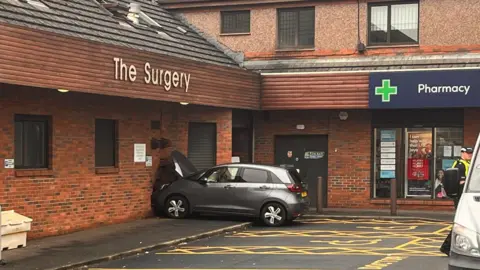 BBC view showing a car crashed into the wall of a GP surgery, next to a boots pharmacy